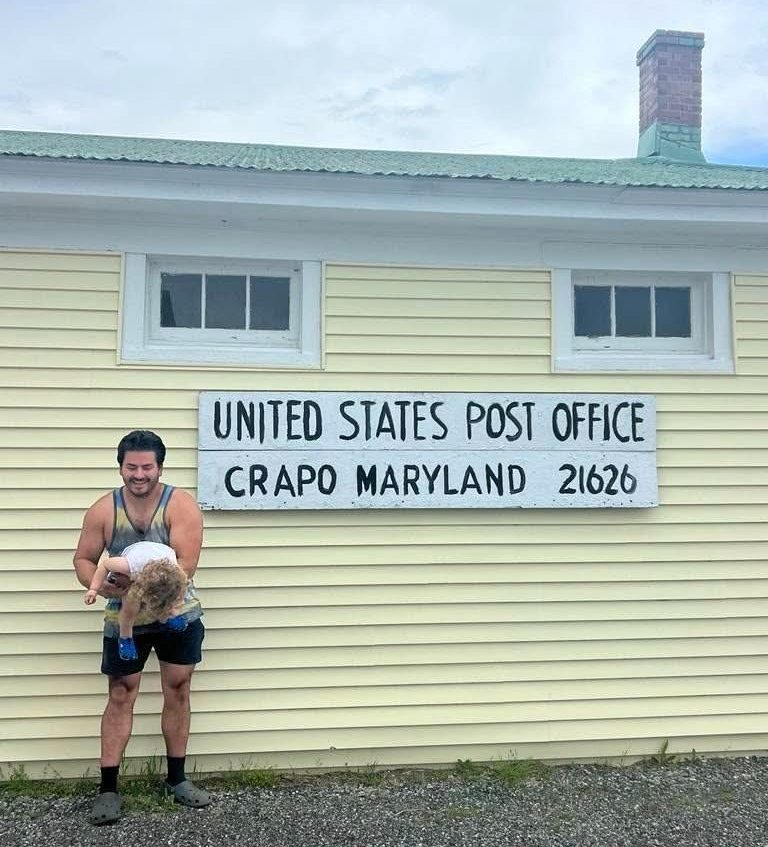 A man holding a playful child in front of a United States Post Office labeled "Crapo Maryland 21626"